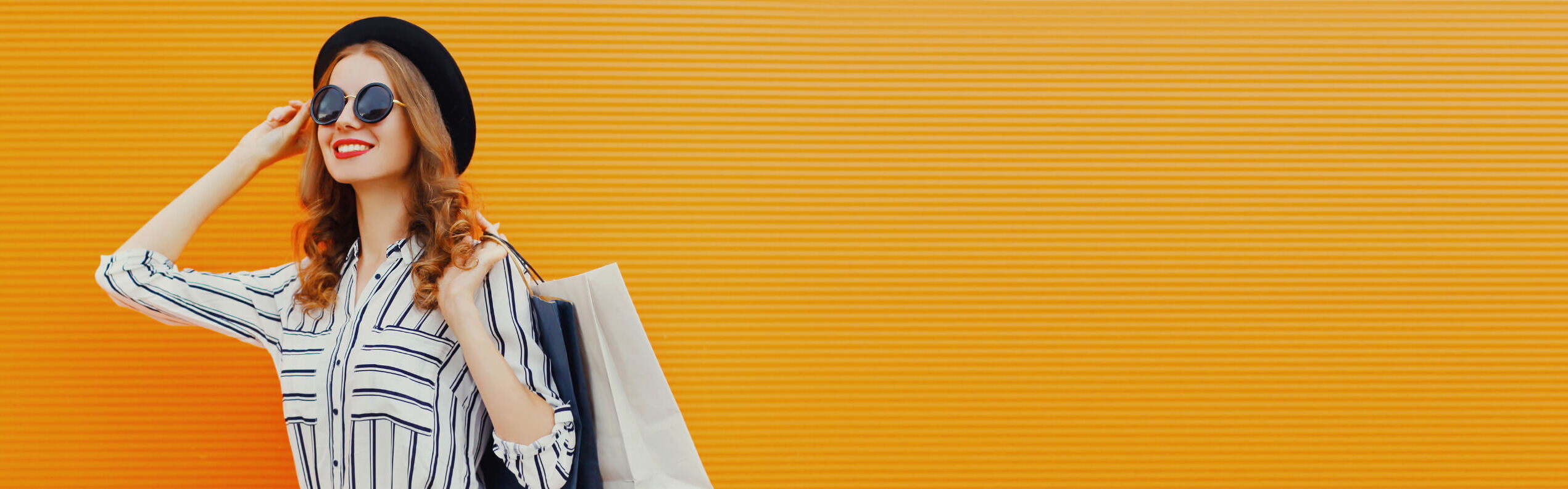 Frau mit Hut und Sonnenbrille in gestreiftem Hemd posiert vor orangefarbenem Hintergrund.